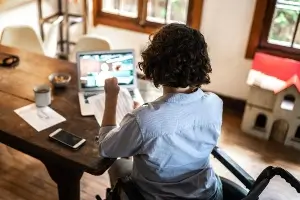image looks over the shoulder of a physically challenged woman sitting in a wheelchair and working on her laptop